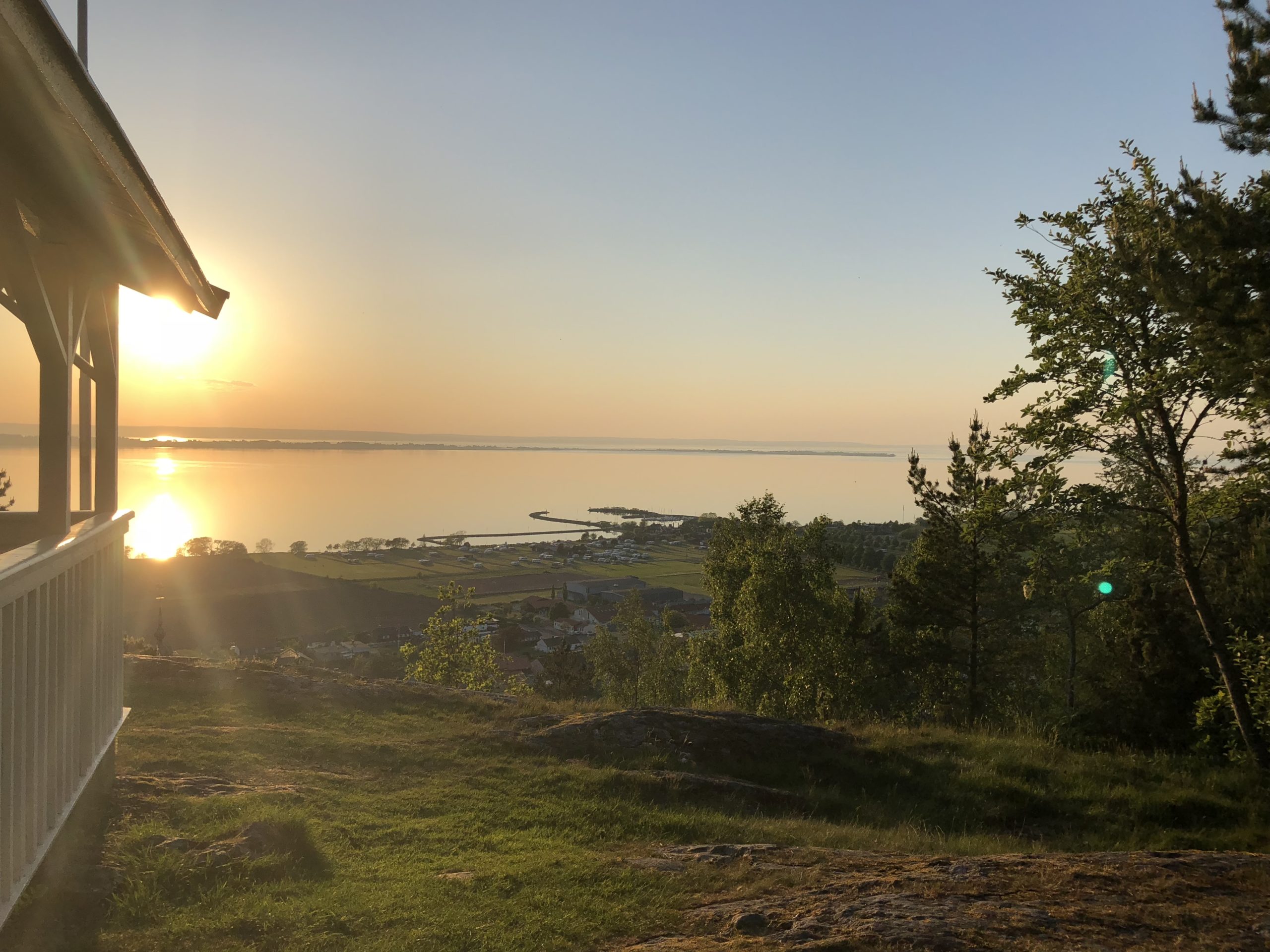 Solnedgång över Vättern sedd från en terrass med gröna träd och gräsmatta i förgrunden på Grenna Hotell i Gränna.