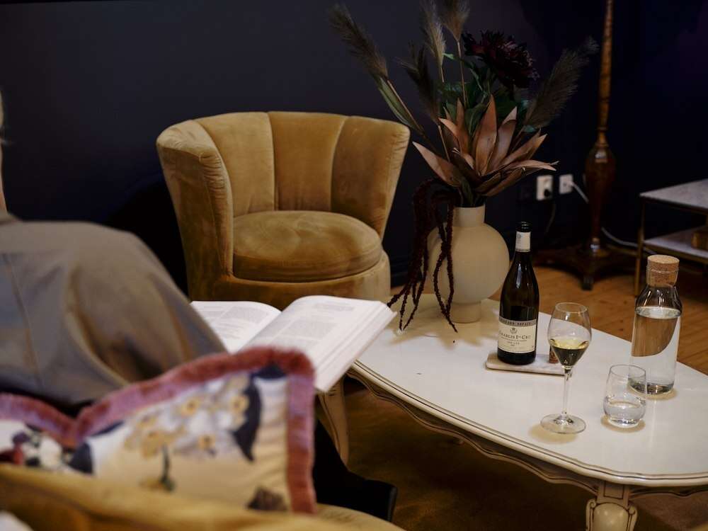 Sittgrupp med senapsgul sammetsfåtölj, soffbord med vinflaska och blomvas vid läsande person på Grenna Hotell