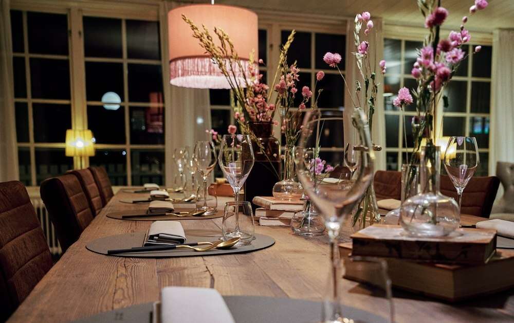 Dukat långbord av trä med vinglas, böcker och rosa blommor i vaser under en taklampa med fransar på Grenna Hotell