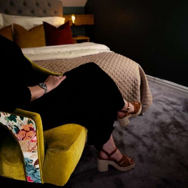 Person sittande i fåtölj framför dubbelsäng i Deluxerum i Annexet på Grenna Hotell