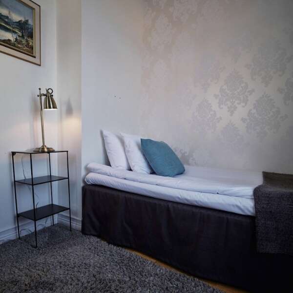 Enkelsäng med blå kudde, bordslampa och ljus mönstrad tapet i Enkelrum på Grenna Hotell