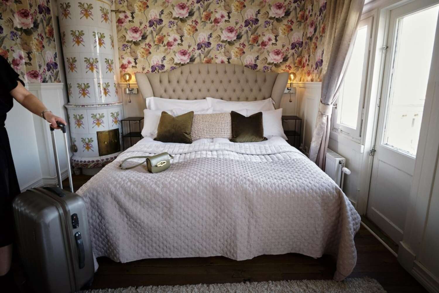 Bäddad dubbelsäng vid kakelugn, blommig tapet och resväska på golvet